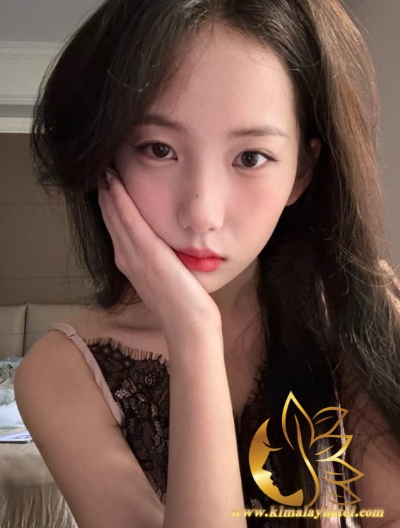 Portrait of a young woman posing with her hand pressed to her cheek, wearing a pink lace camisole indoors.