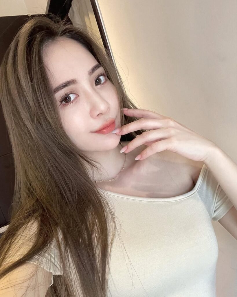 Young woman with long brown hair wearing a light beige off-shoulder top and a delicate necklace, posing with her fingers near her lips.